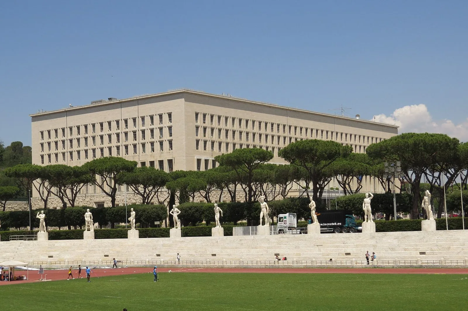 Le gradinate in marmo bianco dello Stadio dei Marmi con le statue di atleti, i pini domestici e il grande Palazzo della Farnesina sotto un cielo azzurro