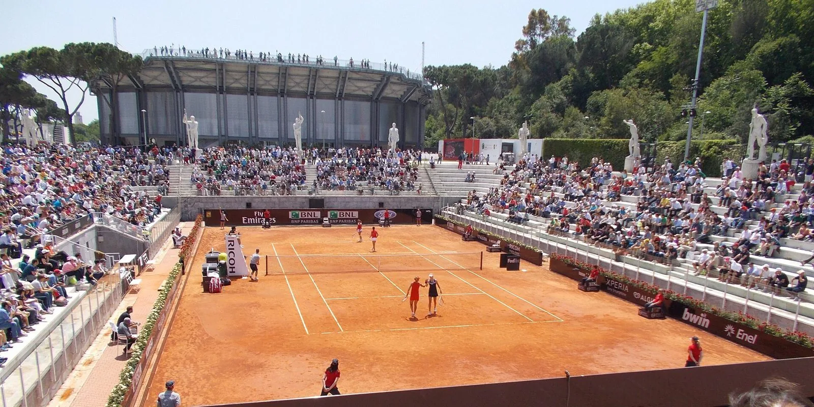 Il campo in terra rossa dello Stadio Nicola Pietrangeli durante una partita di doppio, con le gradinate piene di pubblico, le statue di marmo e il Campo Centrale sullo sfondo