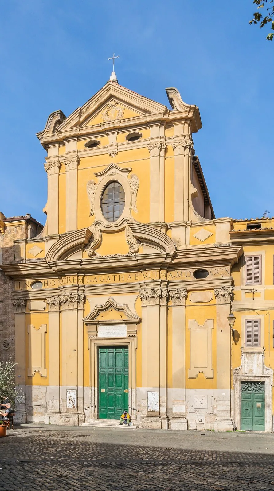La facciata barocca gialla e bianca della chiesa di Sant'Agata in Trastevere, con portone verde, finestra ovale e iscrizione latina, sotto un cielo azzurro