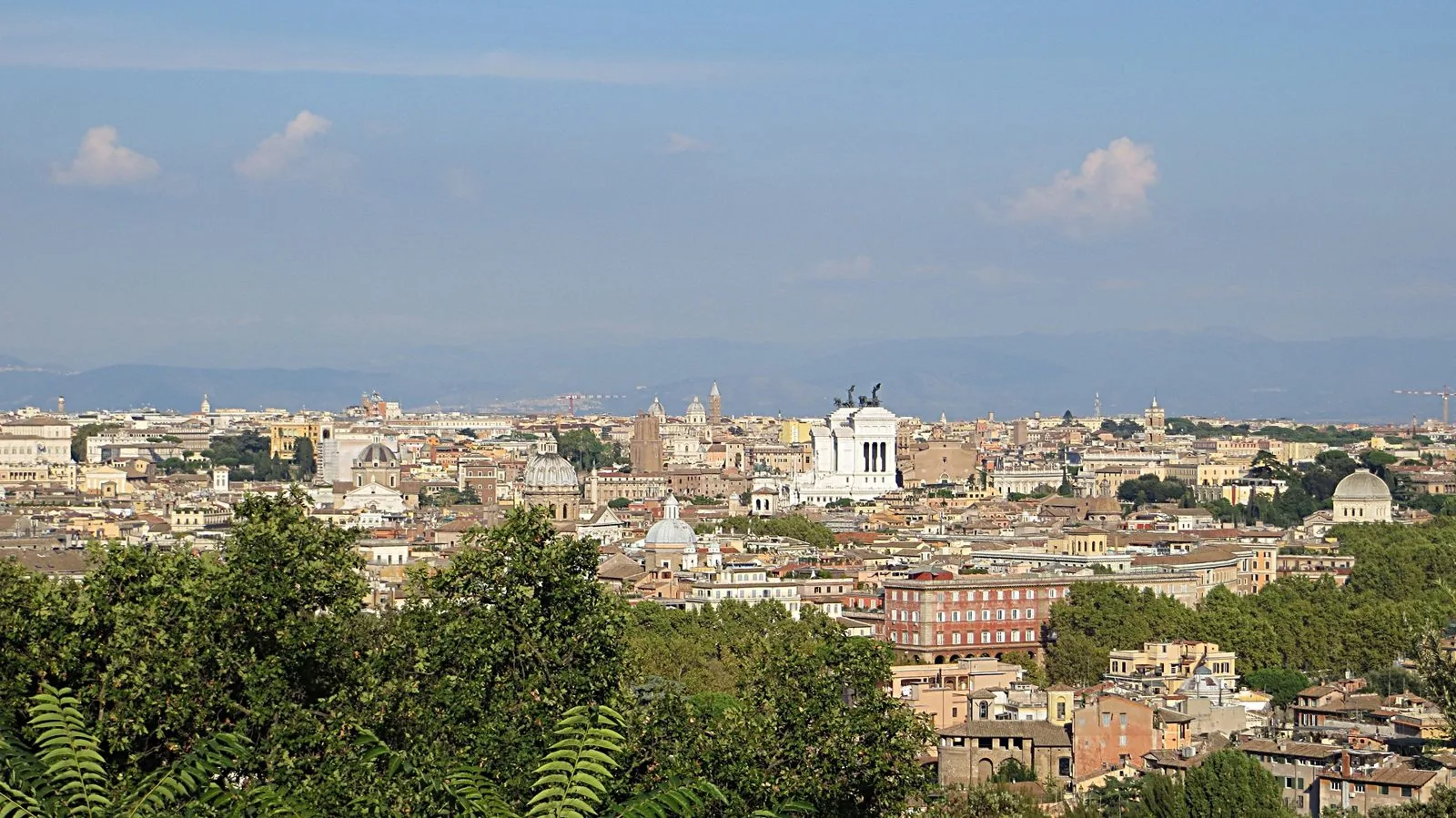 Panorama di Roma dall'alto con tetti, cupole e il bianco Vittoriano al centro, alberi verdi in primo piano e colline sullo sfondo