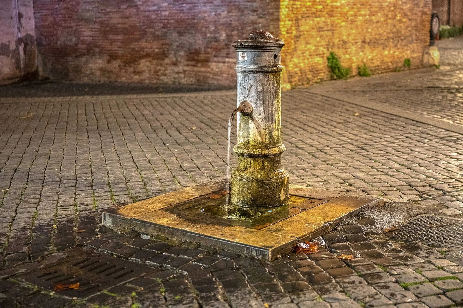 Un nasone, la fontanella pubblica romana in ghisa, con l'acqua che scorre dal beccuccio su una piazza lastricata di sampietrini, di sera